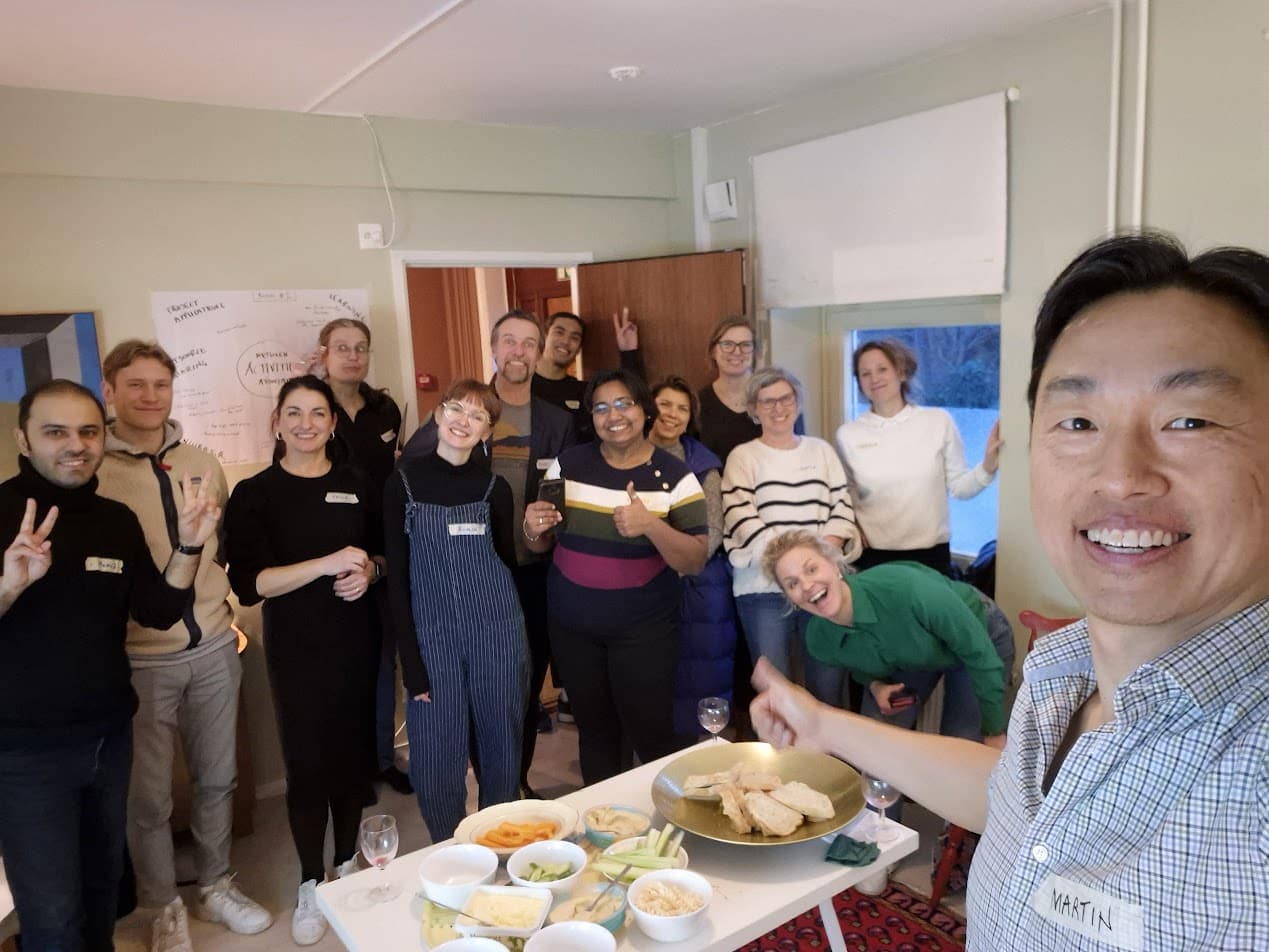 En gruppbild i form av en selfie tagen av ordförande Martin Güll, som syns till höger i förgrunden och gör tummen upp. Bakom honom står ett tiotal glada deltagare och styrelsemedlemmar samlade i ett ljust rum på Ödet. I förgrunden syns ett bord uppdukat med snacks och tilltugg, och i bakgrunden hänger en whiteboard med anteckningar från pre-inkubatorns workshop.