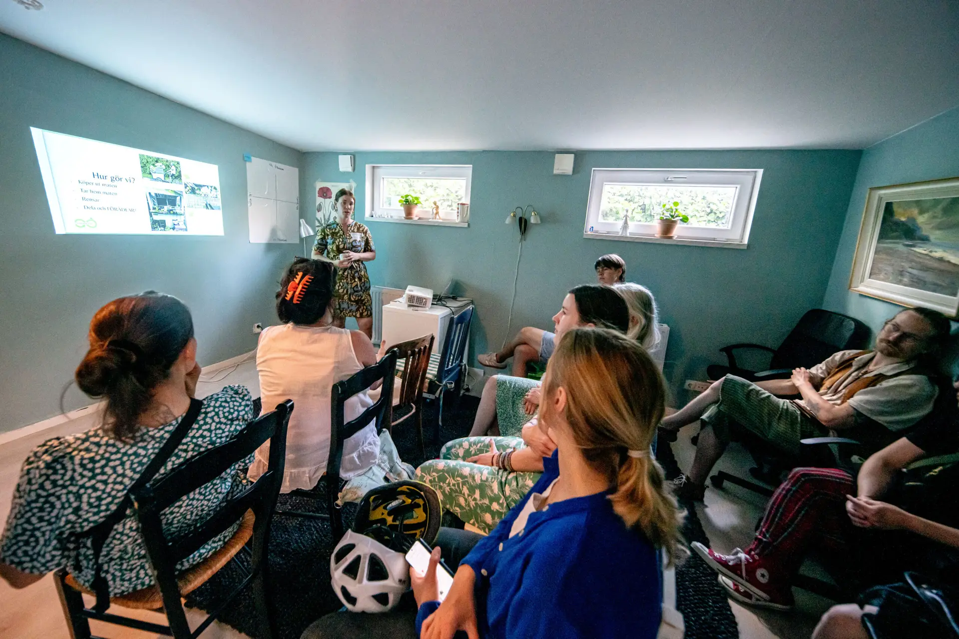 Grupp möts för en presentation i Ödets flexibla och råa möteslokal med blå väggar. En kvinna talar inför sittande deltagare. Foto: Andreas Paulsson.