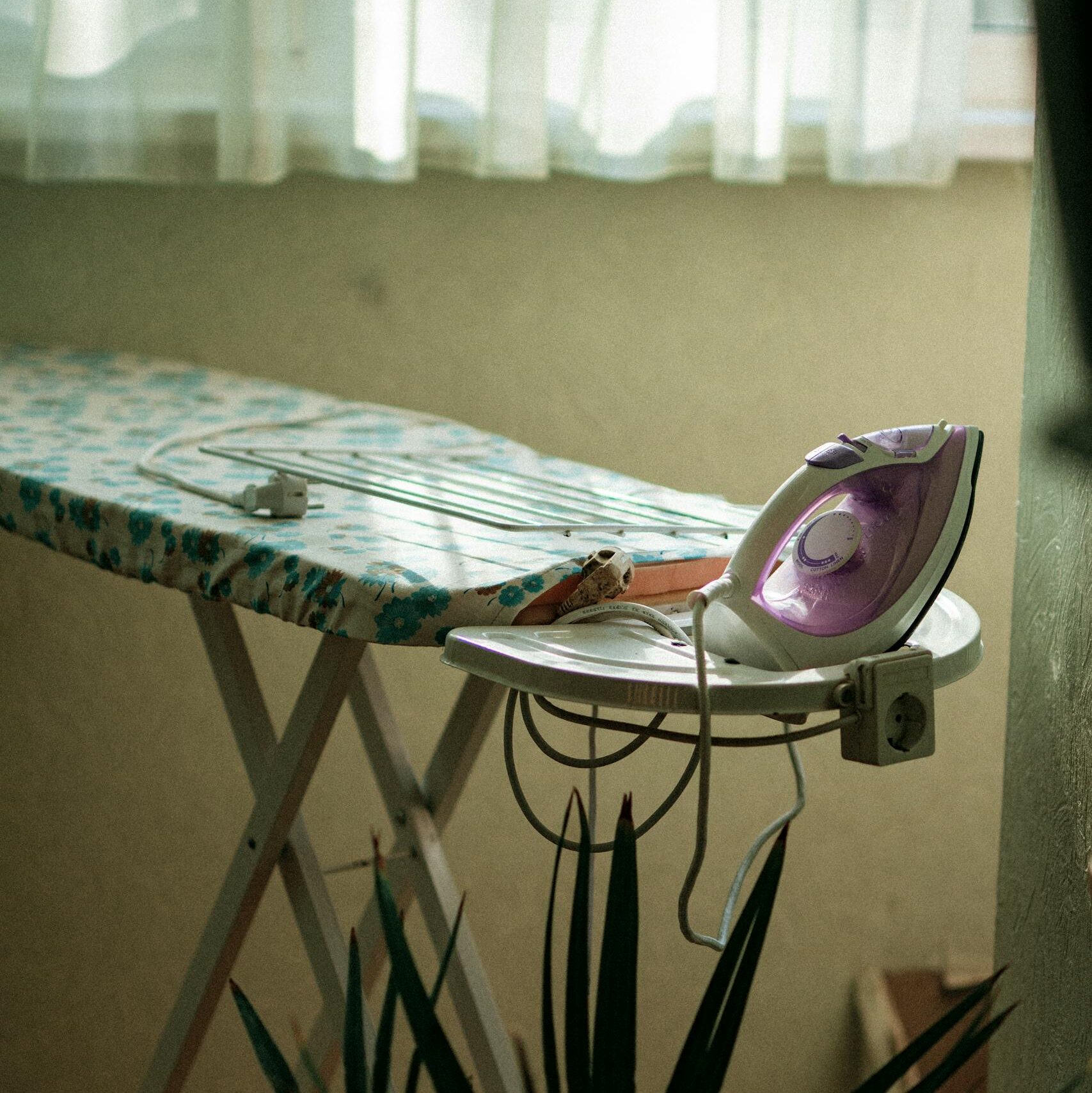 A well-lit ironing board and steam iron near a window with lush green plants.