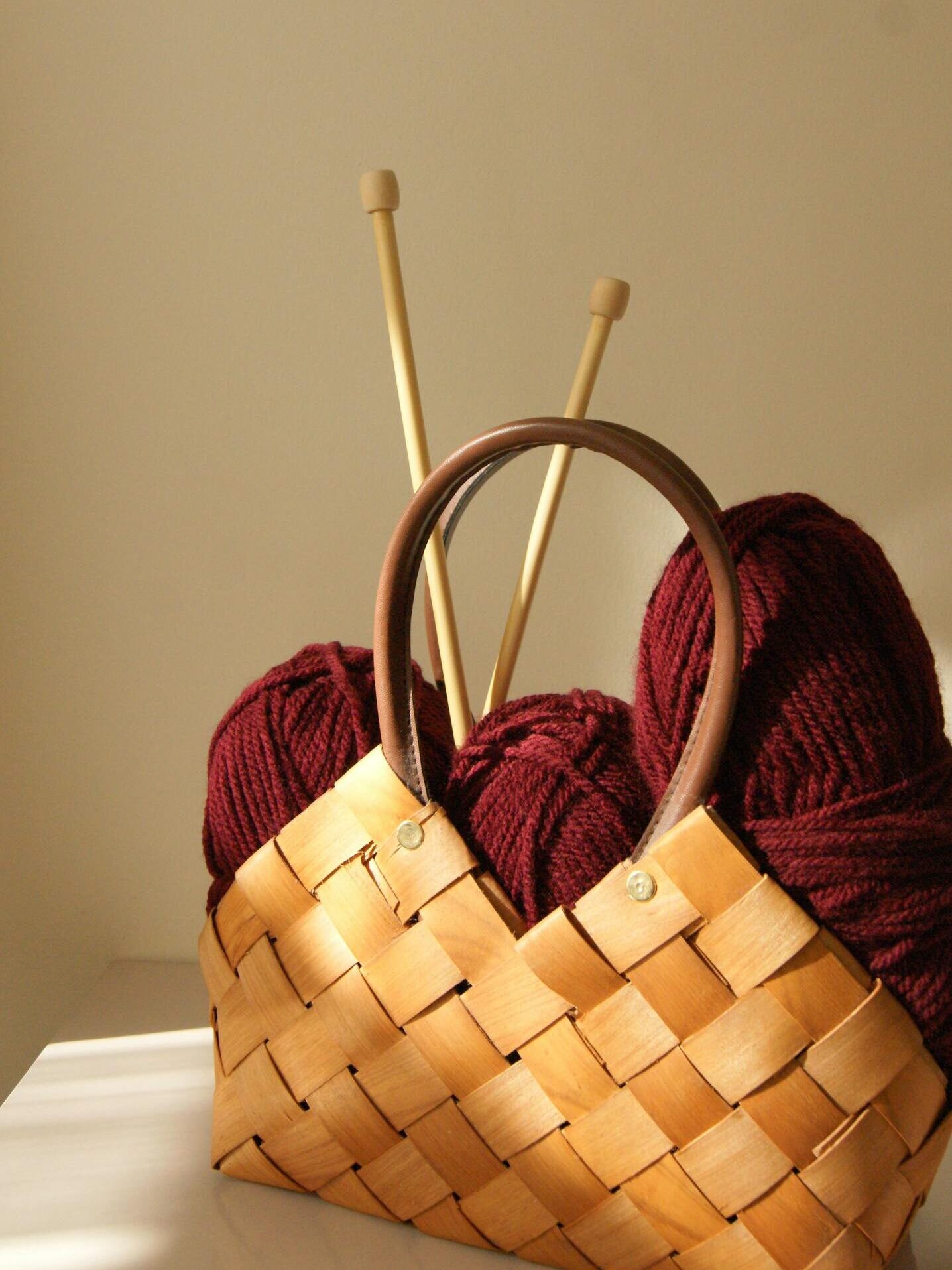 Cozy wicker basket filled with burgundy yarn and wooden knitting needles in warm sunlight.