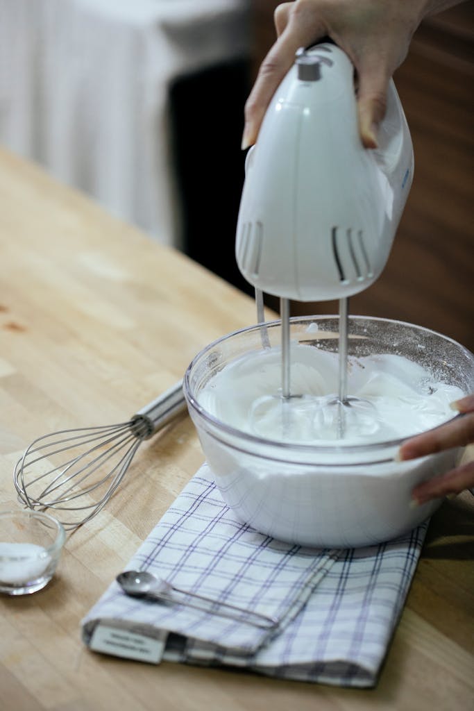 Crop anonymous female chef beating egg whites with modern mixer while cooking pastry in light kitchen